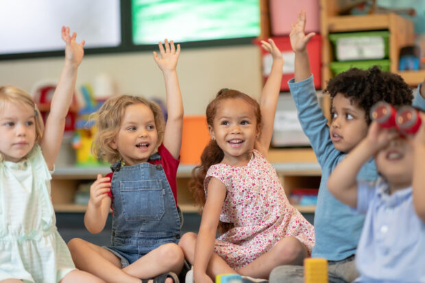 A group of nursery children sat smiling with their hands up.