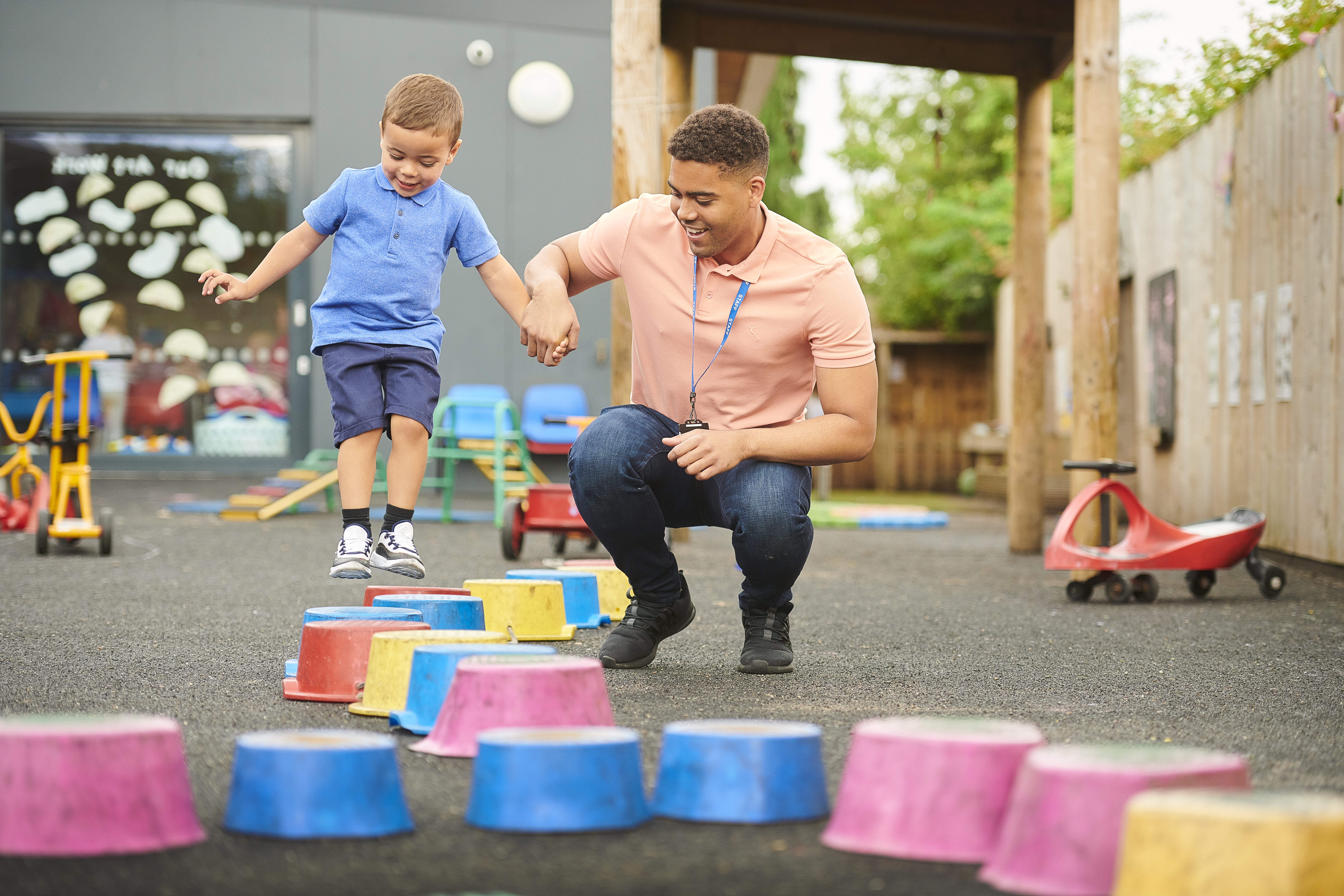 A nursery worker holding hands with a child in a playground. The child is jumping from one pot to another.