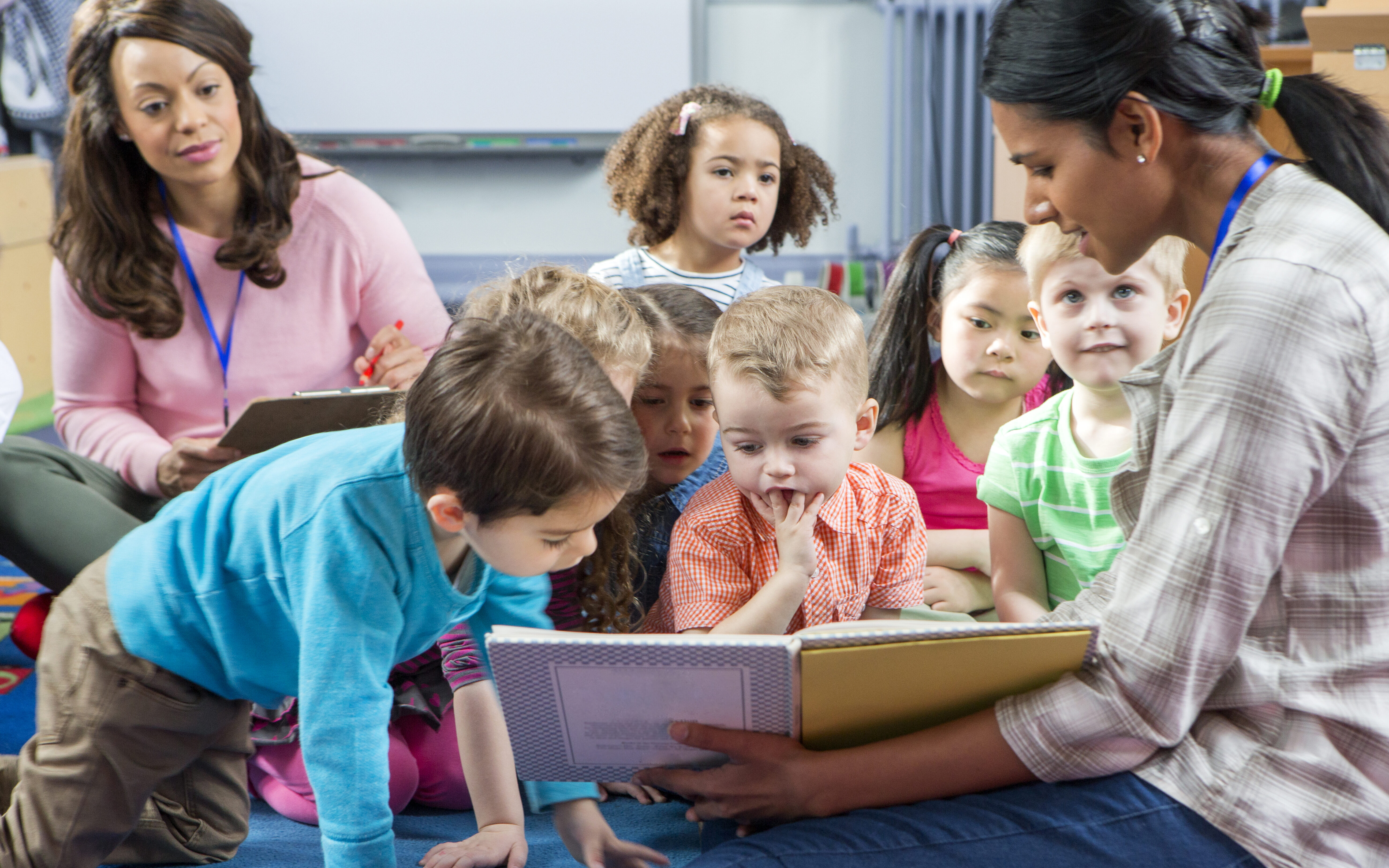 A teacher is giving a lesson to nursery students. They are sitting on the floor looking at a book together